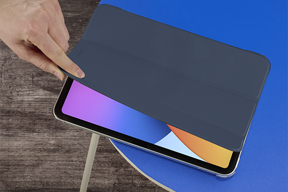The Hama tablet case ‘Fold Clear’ lies on a blue, round tabletop. A hand comes out of the top left-hand corner and opens the lid of the cover, revealing the activated display of the Apple iPad 10.9’.