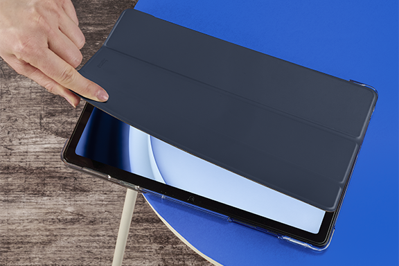 The Hama tablet case ‘Fold Clear’ lies on a blue, round tabletop. A hand comes out of the top left-hand corner and opens the lid of the cover, revealing the activated display of the Samsung Galaxy Tab 9.