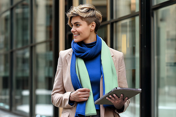 A young woman is out and about on a shopping street, carrying her tablet in the Hama ‘Fold Clear’ tablet sleeve. She is dressed for spring with a beige blazer, blue T-shirt and green and blue scarf.