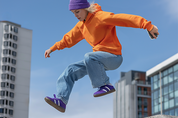 A woman in jeans and an orange jumper jumps over a concrete wall, with skyscrapers and a blue sky in the background