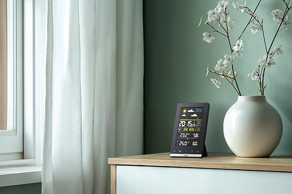 The Hama weather station ‘Jamaika’, with outdoor sensor, radio, alarm clock and indoor climate, stands on a wooden sideboard together with a white vase with dried flowers in it. A window with a white curtain can be seen on the left-hand side
