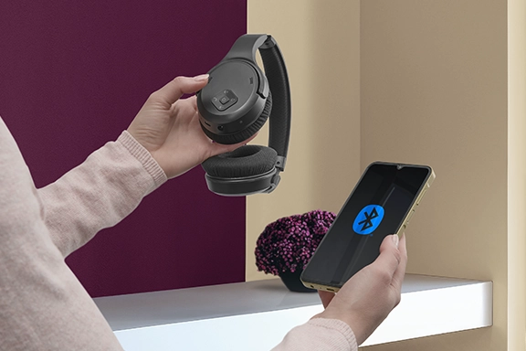A woman holds a smartphone with the Bluetooth logo on the display in her right hand and the Hama over-ear TV headphones in the other.
