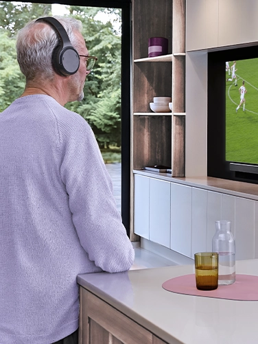 A man is standing in the kitchen watching a football match on television. He is listening to the sound via the Hama TV headphones.