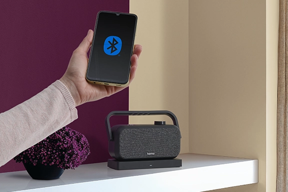 A hand holds a smartphone with a Bluetooth logo on the display in the picture. In the background, the loudspeaker for the television stands on a white sideboard.