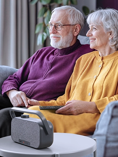 A couple are sitting snuggled up together on the sofa watching TV. The Hama TV speaker is on the table in front of them.