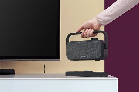 From the right-hand side, a hand extends the portable television loudspeaker into the picture. In the background is a television scene.