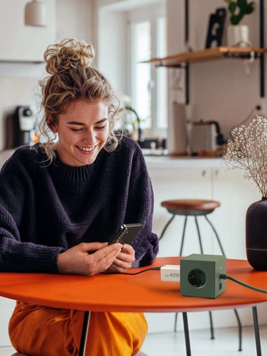 Young woman sitting at a dining table and operating her smartphone while it is being charged with the Hama fast charger, 45W