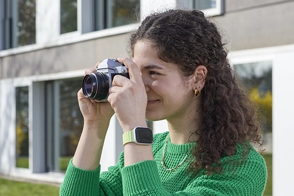 A young woman in a green knitted jumper wears an Apple Watch together with the Hama ‘Fantastic Feel’ magnetic wristband and photographs her surroundings with a retro camera.