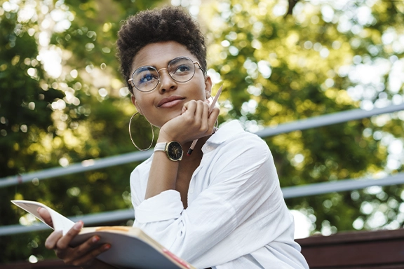 A woman in a light white blouse is sitting on a bench in the countryside. On her wrist she is wearing the Hama Smartwatch "7000" with AMOLED 43 mm display and phone function