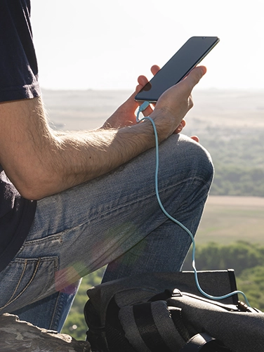 Man sits on a hill while hiking and charges his smartphone with the Hama Power Pack "PD10-HD"