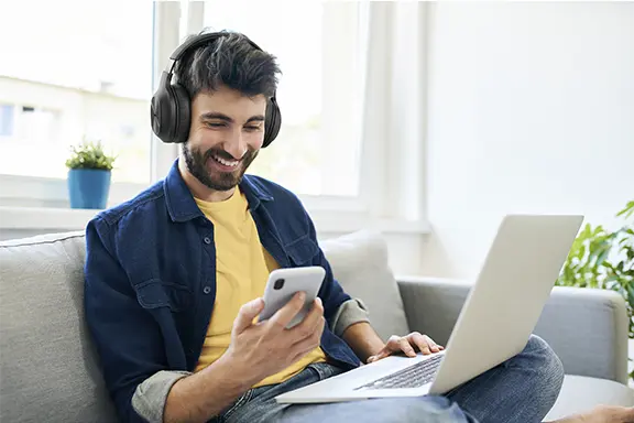 A man wearing headphones is sitting on a sofa, smiling and looking at his mobile phone; he has a laptop on his lap, with windows and plants in the background