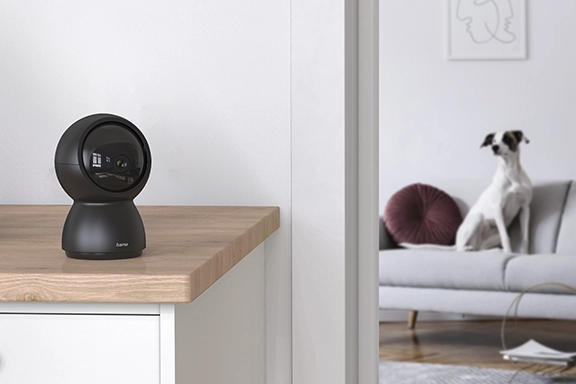The swivelling Hama surveillance camera with WLAN and motion detector for indoors stands in the foreground on a wooden sideboard. A dog sits on the sofa in the background.
