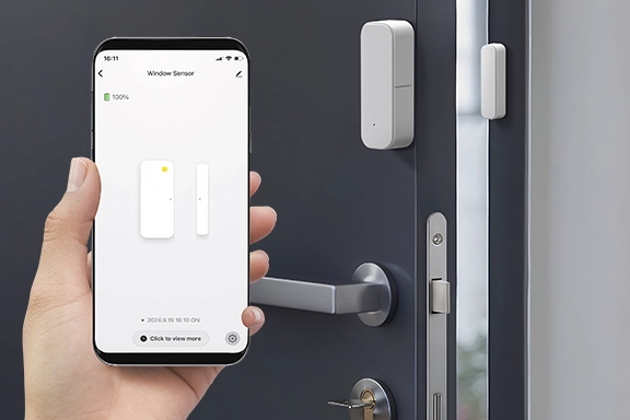 In the foreground is a hand holding a smartphone on which the Hama Smart Home app is open. The smart door & window contact can be controlled here. In the background you can see a dark grey front door to which the door contact is attached.
