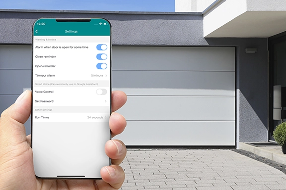 In the foreground, a hand holds a smartphone with the Smart Home app open. In the background you can see a driveway with a closed garage door.