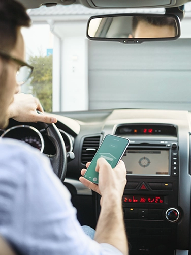 A man sits in his car in front of his garage. With his smartphone in his hand, he can open the garage automatically via the Smart Home app.