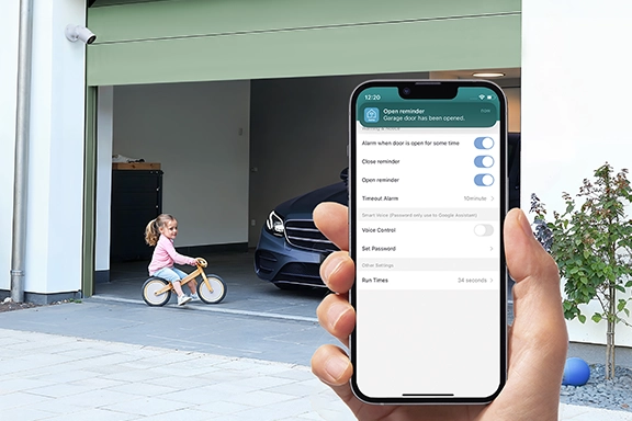 In the foreground, a hand holds a smartphone with the Smart Home app open. In the background you can see a driveway with an open garage door.