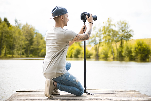 A young man kneels on a jetty by the lake and photographs the scene with his SLR camera, which is mounted on the Hama Monostand "Smooth"