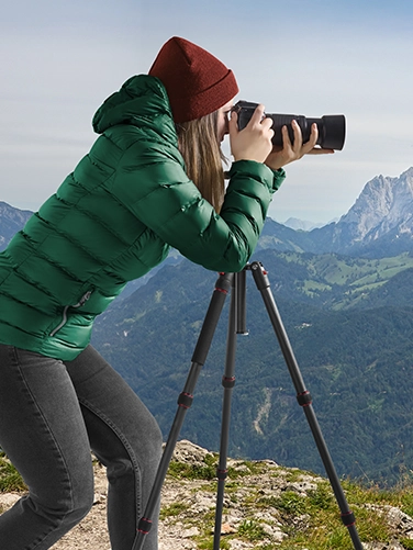 Young woman is dressed in a jacket and cap on a hill in the mountains and takes pictures with an SLR camera mounted on the Hama tripod "Ramble II Duo Carbon"